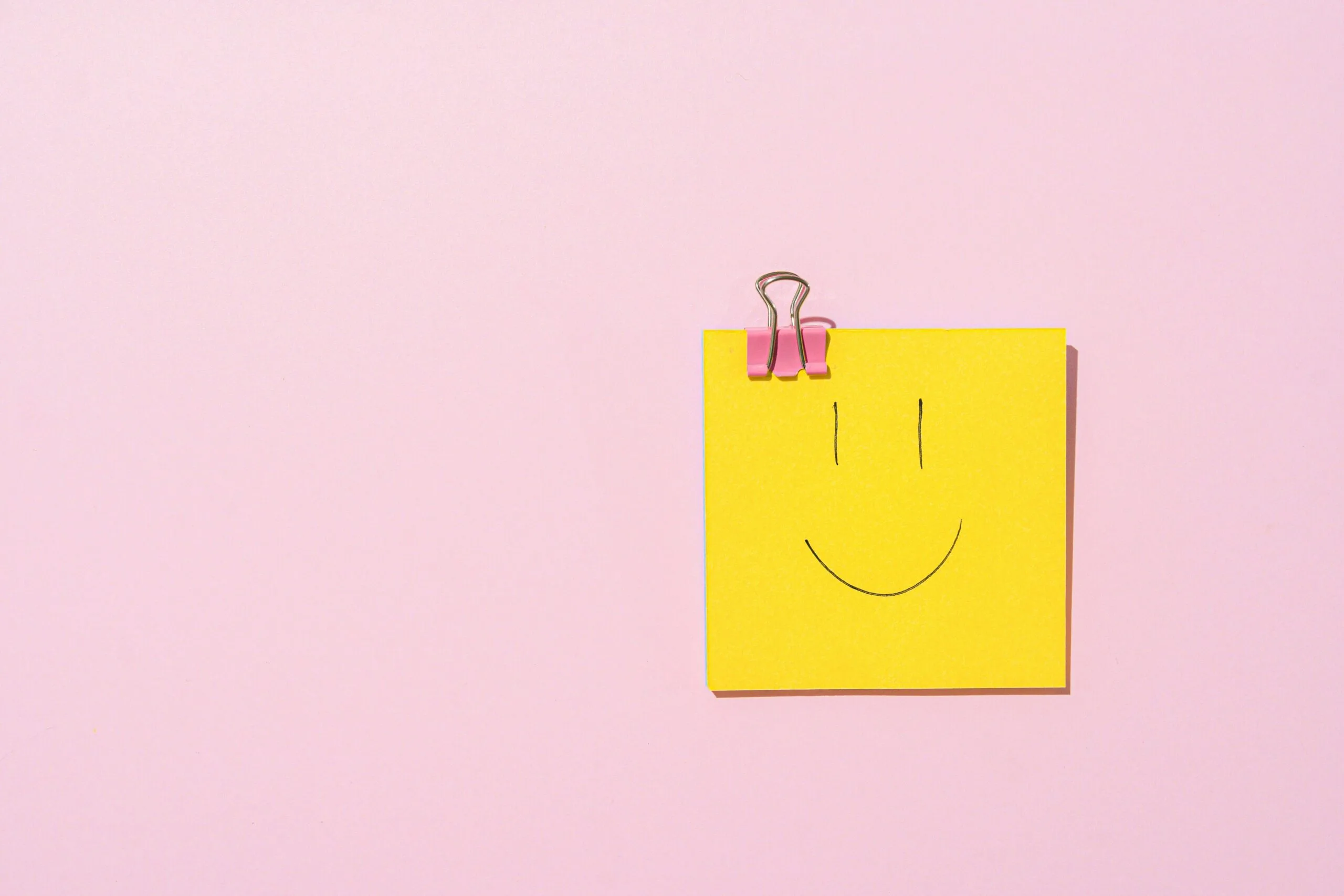 Yellow sticky note with a cute smiley face drawn on a sticky not and clipped to a pink surface